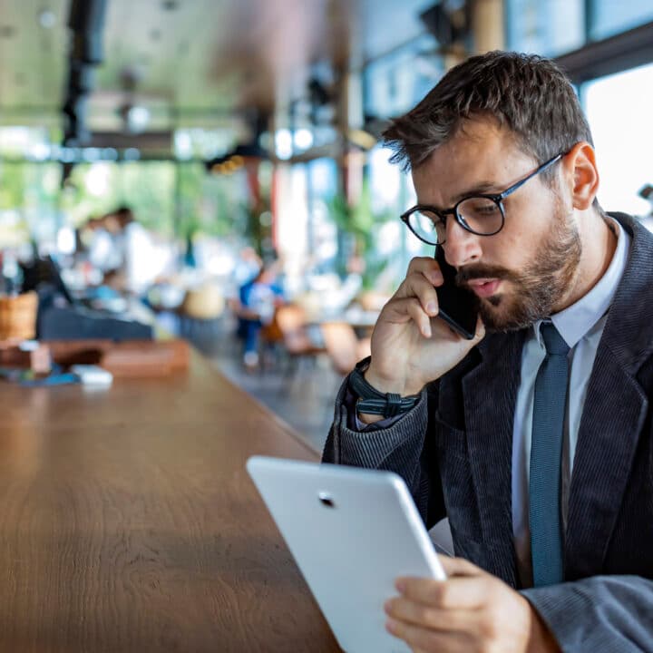 Happy and Successful Male Business Person with Beard, Dressed in Casual Suit is Communicating over a Mobile Phone and Drinking Coffee in Modern Restaurant. A Modern and Handsome Businessman is Sitting in City Café, Enjoying in Hot Cappuccino and Talking about Business with his Colleagues.