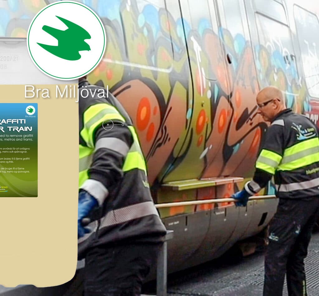 Bilden visar två personer som arbetar med att ta bort graffiti från ett tåg. De använder en produkt som marknadsförs som miljövänlig, vilket framgår av texten &#34;Bra Miljöval&#34; och etiketten på produkten. Tåget är täckt med färgglad graffiti.
