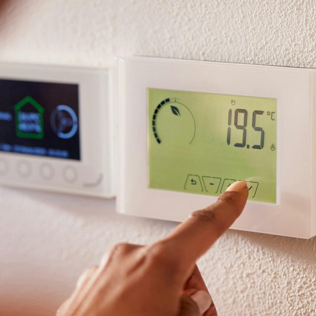 Bilden visar en persons hand som justerar en digital termostat upphängd på en vägg. Termostaten visar en temperatur på 19.5°C. Det finns knappar på termostaten som personen verkar trycka på för att antagligen ändra inställningarna. Bakom termostaten syns en annan enhet monterad på väggen, men det är oklart vad det är eftersom den är suddig och endast delvis synlig. Väggen i bakgrunden verkar ha en ljus färgton.