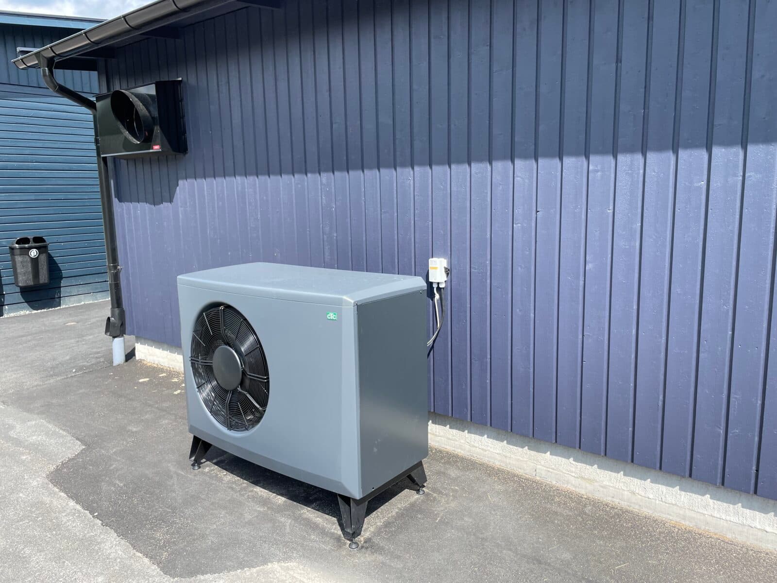 Luftvärmepump placerad utomhus mot en blå vägg. Pumpen är grå och ansluten till en strömkälla.